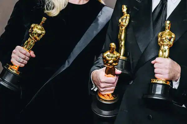 Oscars -  Close-up of two individuals holding their golden Oscar statuettes, one in a black gown and the other in a tuxedo, both proudly displaying their awards.